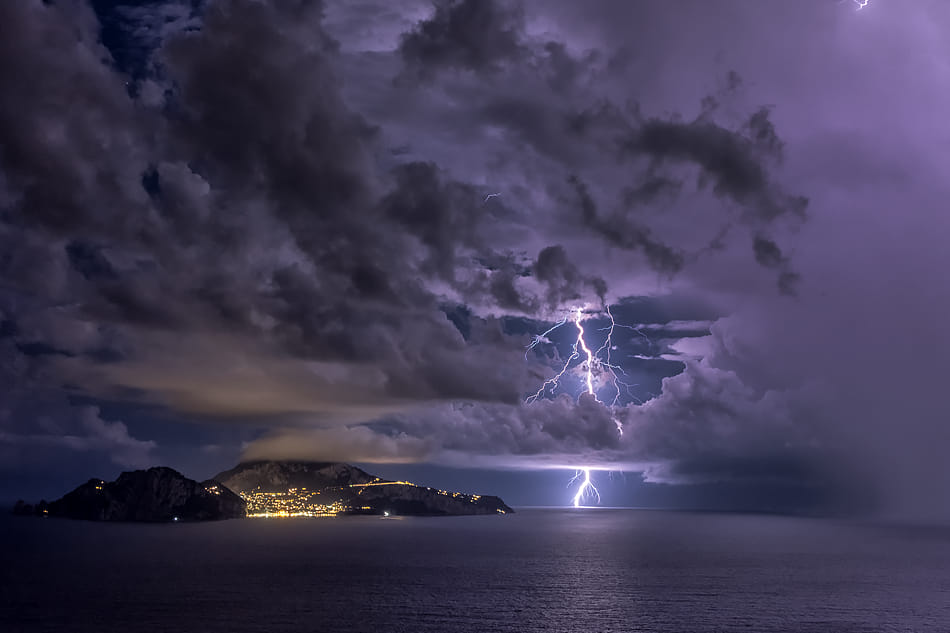Tempesta notturna sul mare: fulmine centrale e costa illuminata, intensità e profondità visiva